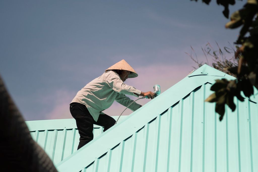 Roof painter spraying a fresh pale-teal coat onto a corrugated metal roof